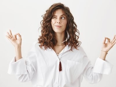 Studio portrait of charming positive caucasian female businesswoman trying to relax while meditating, standing with lifted hands and zen signs over gray background, peeking and looking aside playfully.